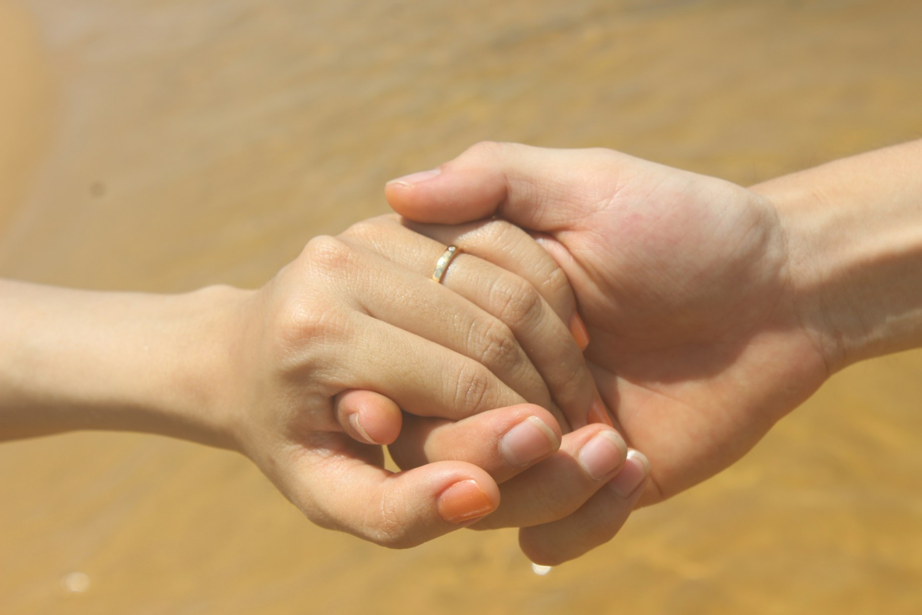 Two hands clasped together, both wearing rings, against a warm sandy background